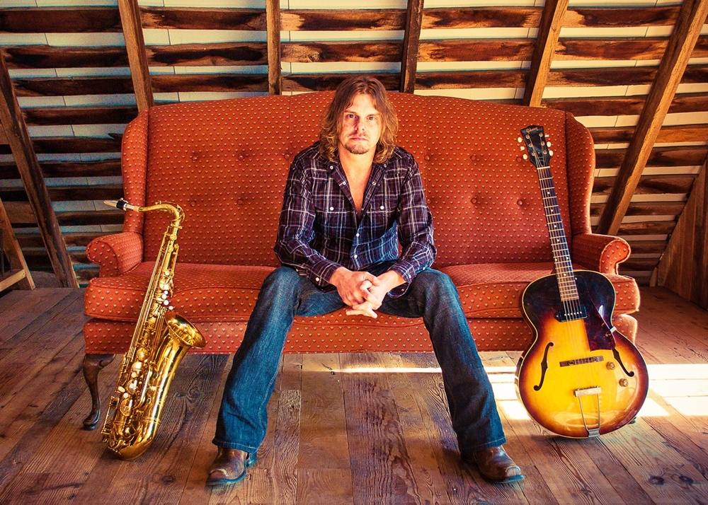 Ken Wenzel sitting on a small sofa with a Saxophone and guitar