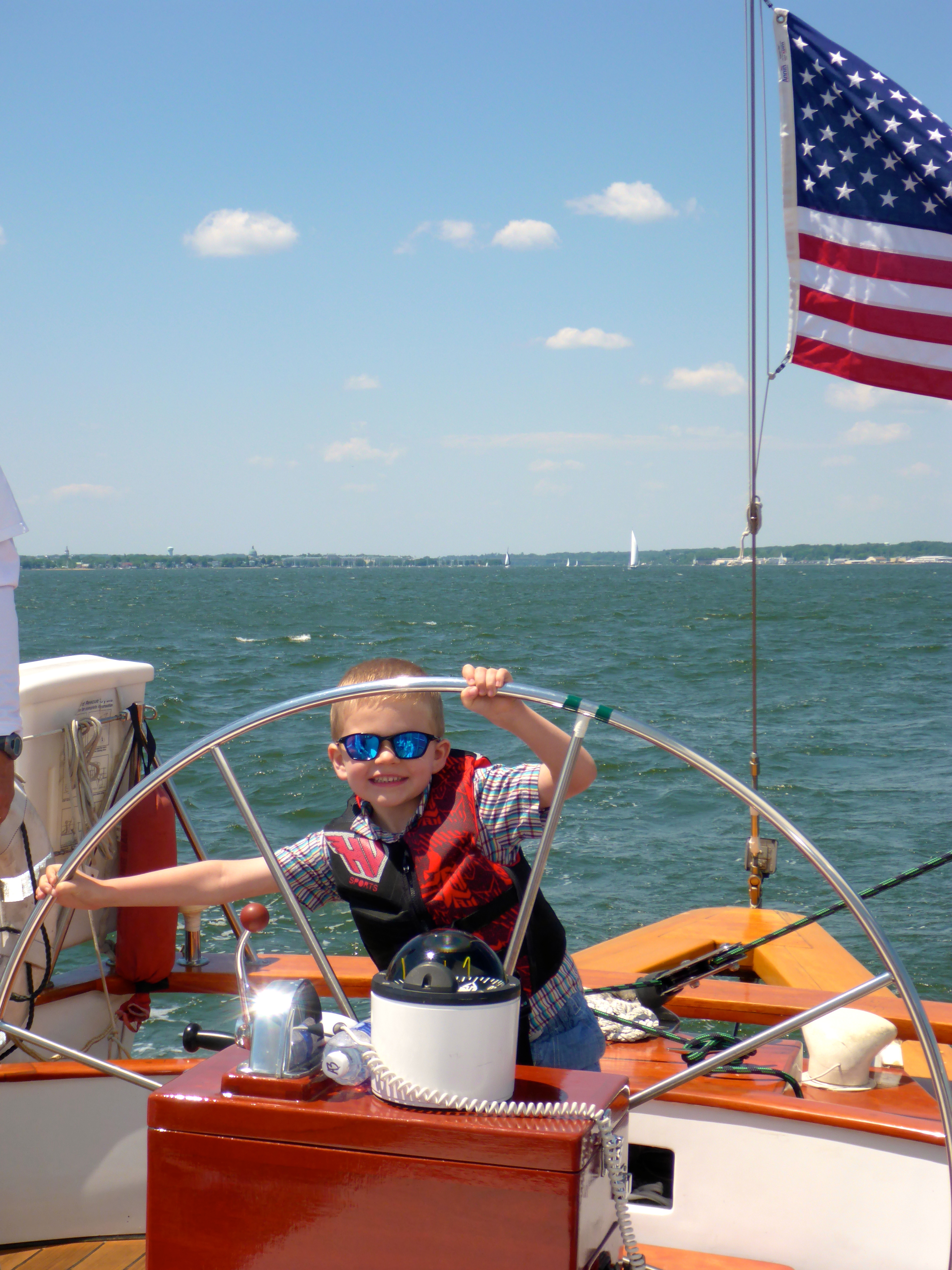 Small boy with sunglasses and life vest smiling as he peaks through the wheel of the schooner