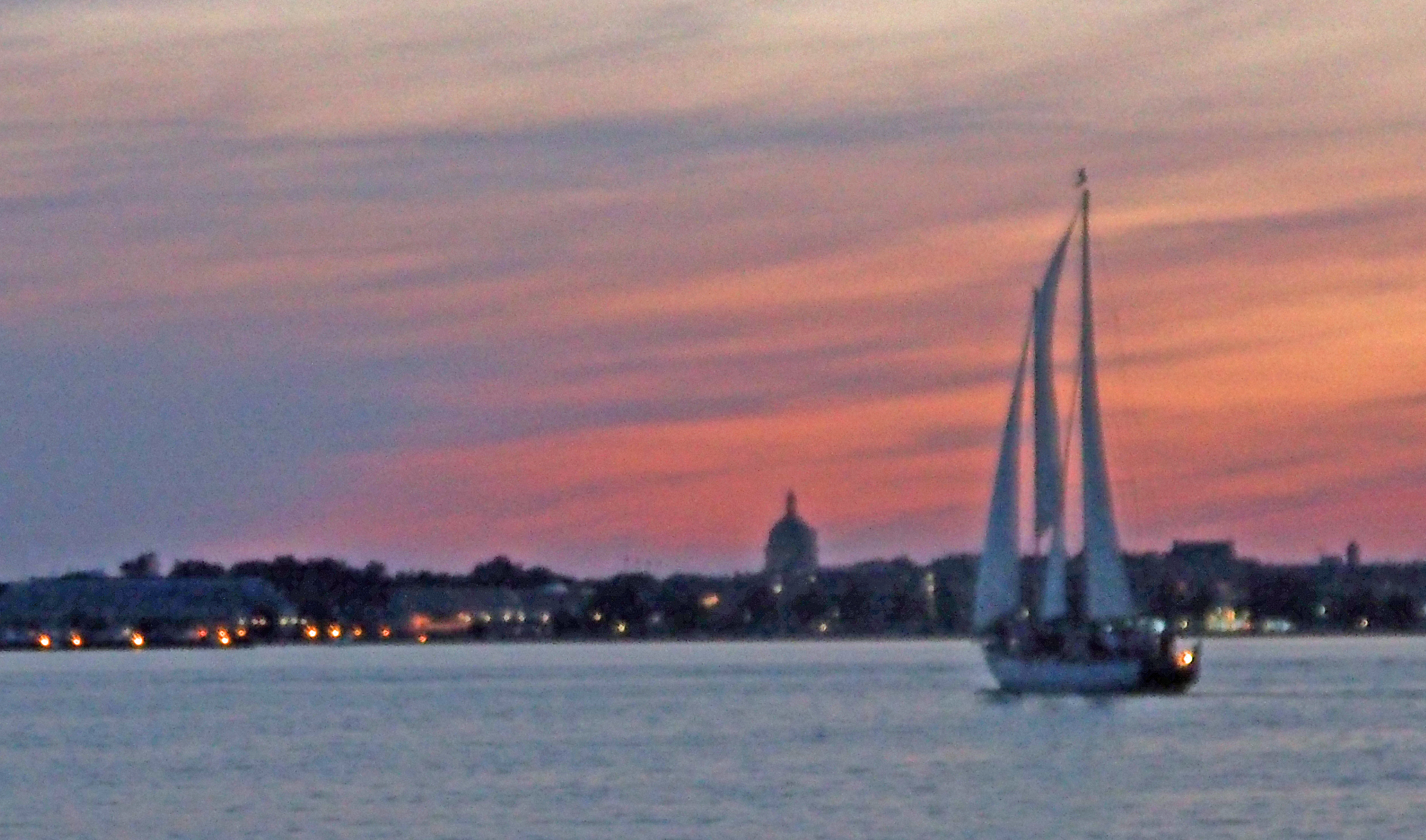 USNA in silhouette against a pinked streaked sky with sailboat on water