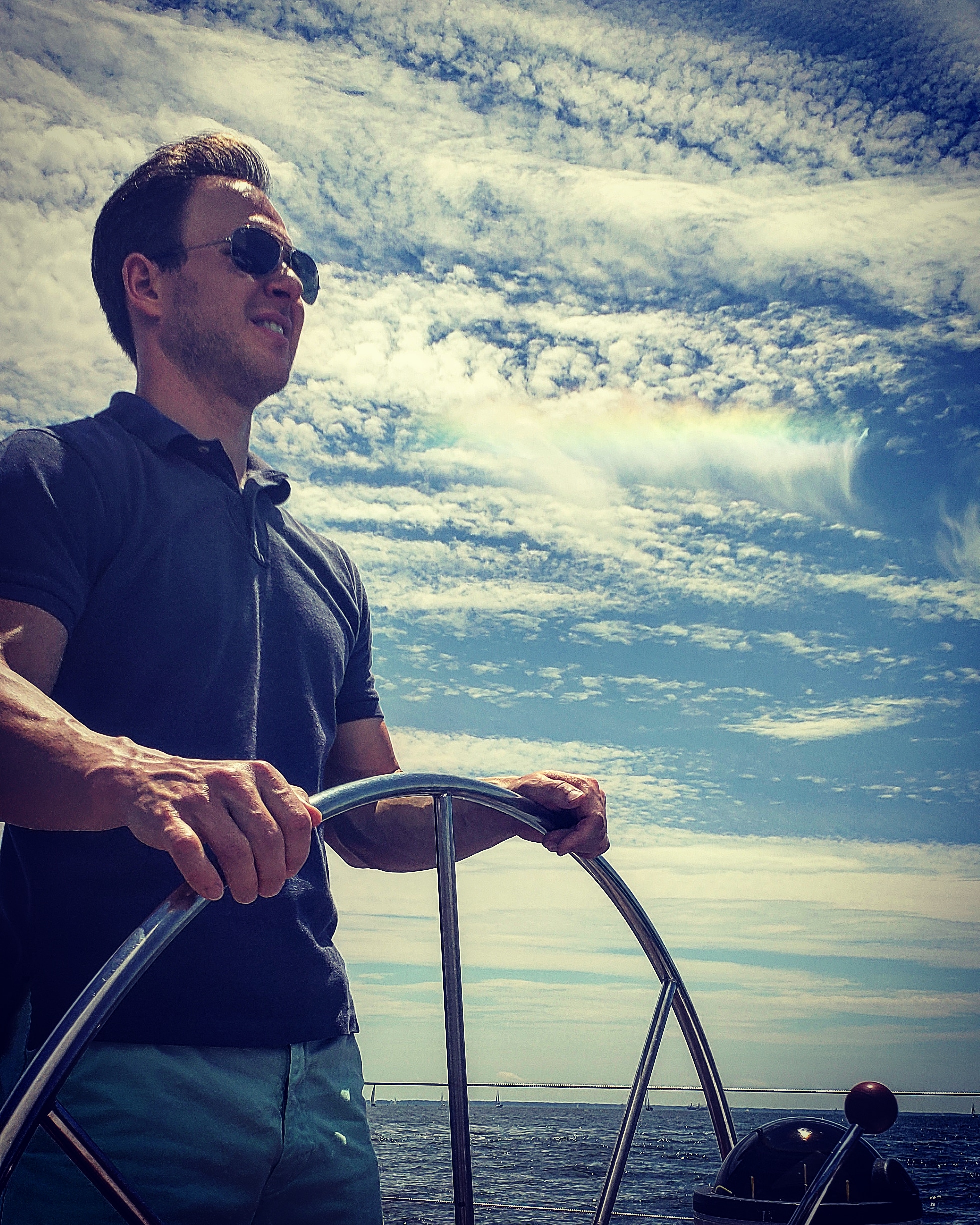 Man steering boat with blue skies and rainbow clouds behind him